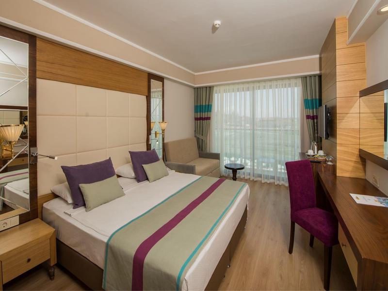 Modernes Hotelzimmer mit Doppelbett, violetten Akzenten, Schreibtisch, Sessel und großen Fenstern mit Vorhängen.