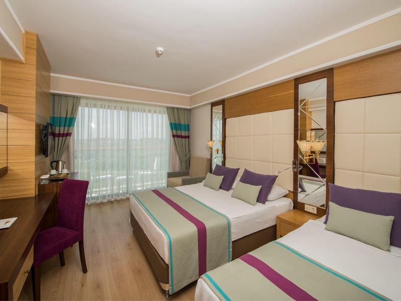 Helles Hotelzimmer mit zwei Betten, Holzmöbeln, lila Stühlen und großem Fenster mit Gardinen.