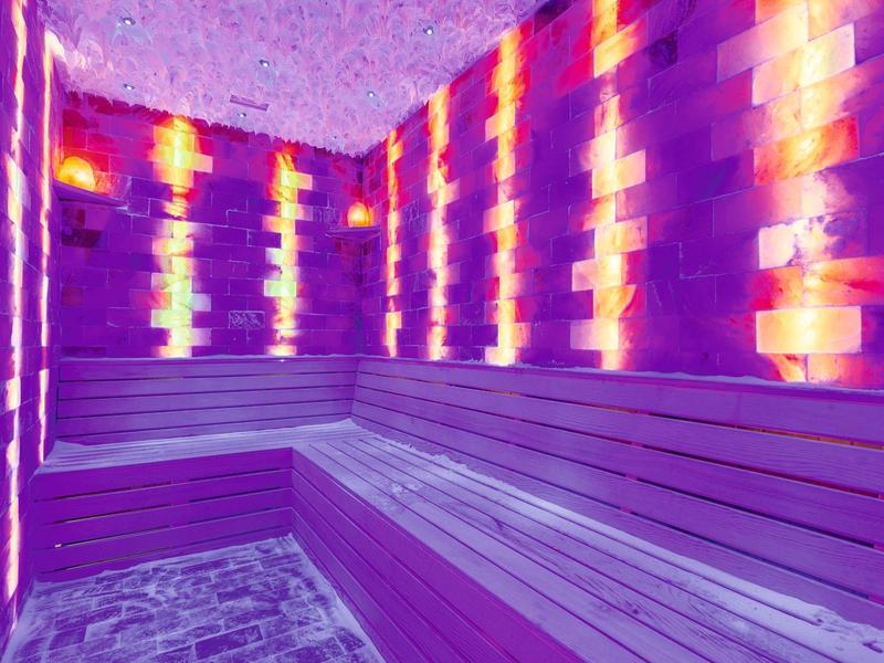 Moderner Sauna-Innenraum mit Holzbank, Ziegelwänden und atmosphärischer lila-oranger Beleuchtung.