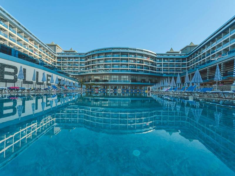 Großes Hotel mit drei Etagen und großem, klarem Außenpool in hellem Blau bei Tageslicht.