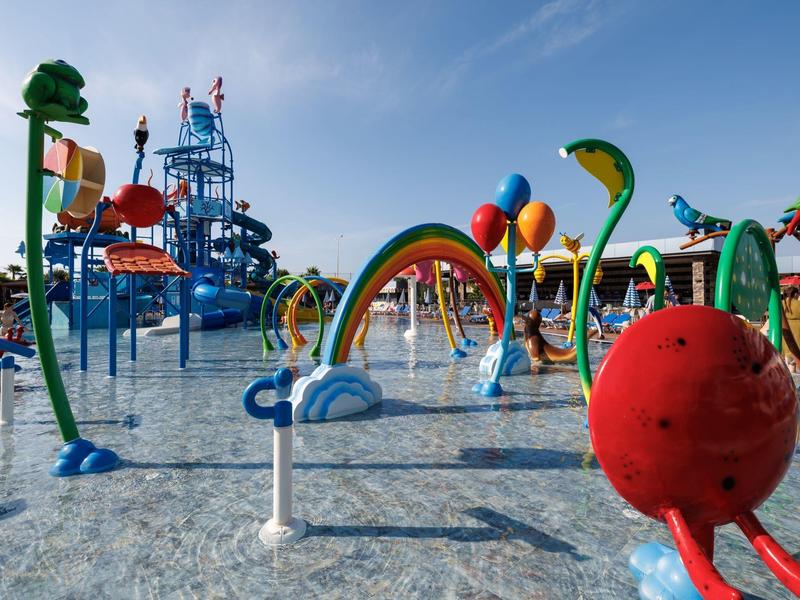 Buntes Wasserspielplatz mit Rutschen, Wasserkanonen und Regenbogenbögen unter blauem Himmel.