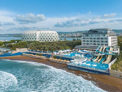 Modernes Hotel mit Rutschen am Meer, Strand und rundem Gebäude im Hintergrund unter blauem Himmel.