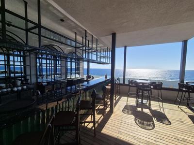 Moderner Innenraum mit Holzboden, Stühlen und Tischen, großer Glasfront mit Meerblick bei Sonnenlicht.
