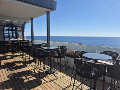 Terrasse mit schwarzen Stühlen und Tischen vor blauem Meer und klarem Himmel.