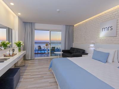 Modernes Hotelzimmer mit Doppelbett, Balkon, Sofa und heller, minimalistischer Einrichtung bei Sonnenuntergang.