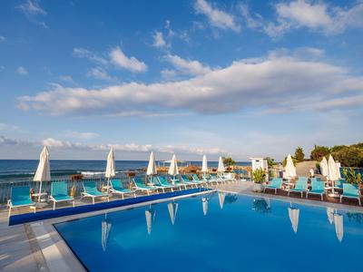 Velký bazén s bílými slunečníky a lehátky s výhledem na moře za jasné oblohy.