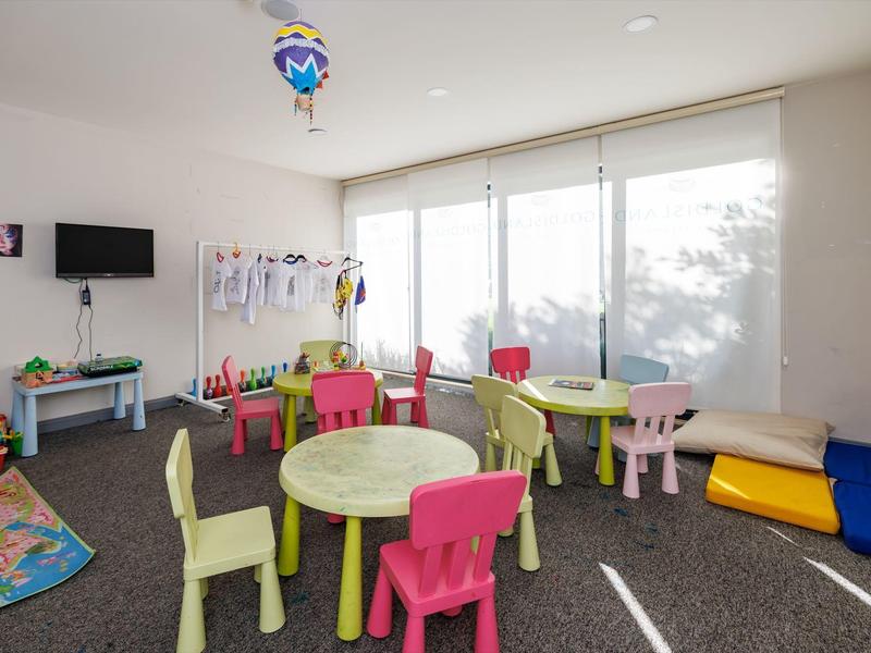 Světlá herna pro děti s barevnými stoly a židlemi, hračkami a velkými okny.