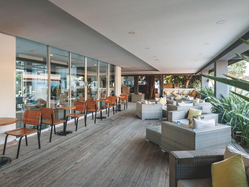 Moderní balkonová plocha s dřevěnou podlahou, sezeními a stoly vedle velkých skleněných oken.