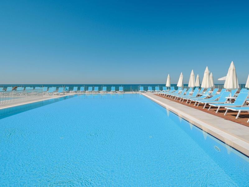 Velký bazén s modrou vodou a lehátky u moře pod jasnou oblohou.