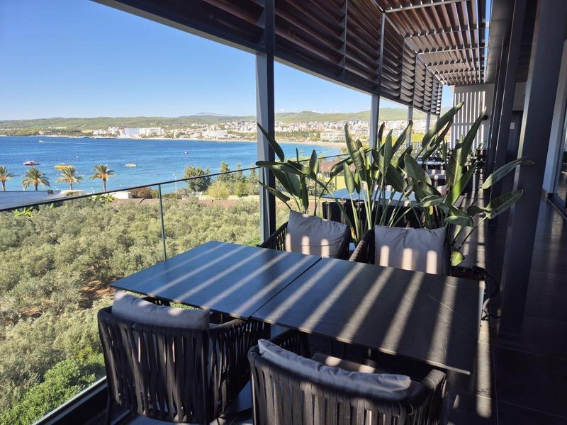 Balkon s stolem a židlemi, výhled na moře a palmy se slunečními stíny.