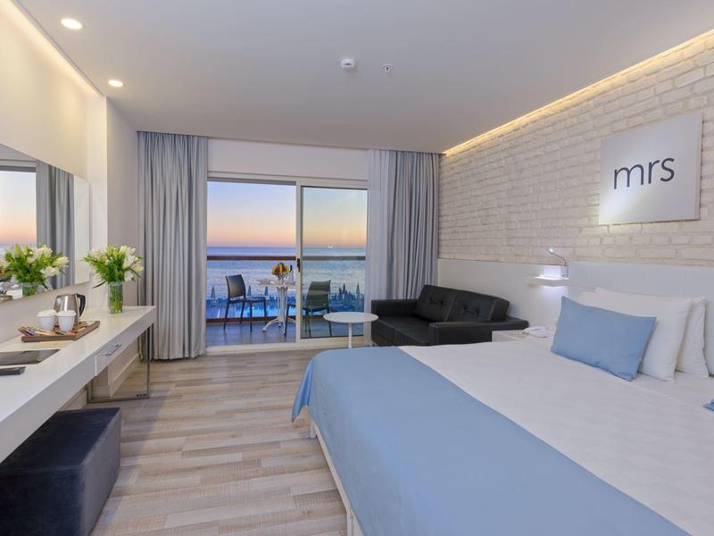 Moderní hotelový pokoj s manželskou postelí, pohovkou, balkonem a výhledem na moře při západu slunce.