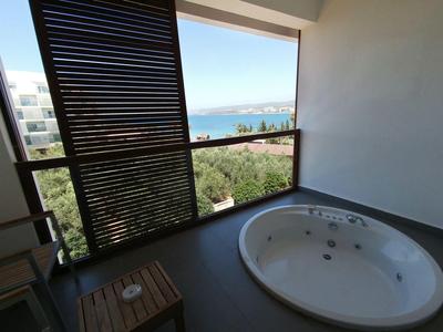 Balkon s vířivkou, výhled na moře a zeleň, dvě dřevěná křesla a malý stůl.