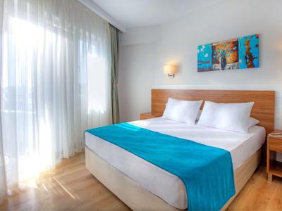 Hell eingerichtetes Schlafzimmer mit großem Bett, blauer Tagesdecke und bodentiefen Fenstern.