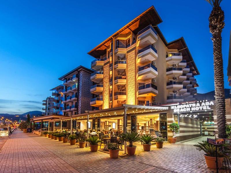 Modernes Hotel mit beleuchteten Balkonen, Außenrestaurant und Palmen bei Abenddämmerung.