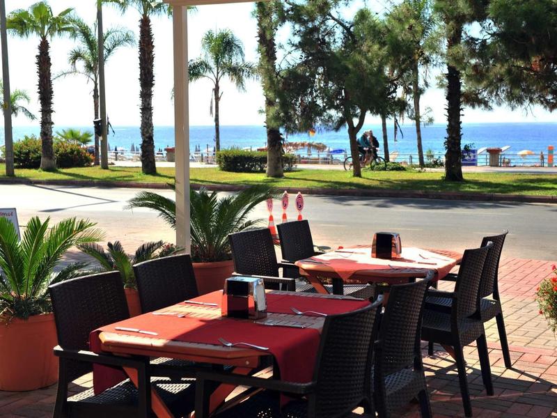 Freiluftrestaurant mit roten Tischen und schwarzen Stühlen, Palmen und Meer im Hintergrund.