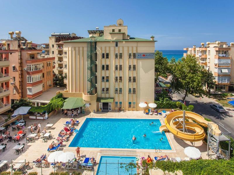Hotel with large outdoor pool, water slide, and sun loungers surrounded by multistory buildings by the sea.