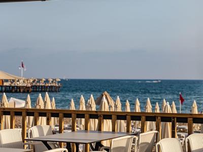 Freie Sitzplätze mit weißen Stühlen und Tischen, Sonnensegel darüber, verlassener Strand mit geschlossenen Schirmen.