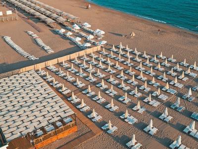 Strand mit gleichmäßig angeordneten weißen Liegen und Sonnenschirmen neben blauem Meer.