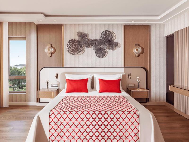 Modern deluxe room with double bed, red accents, wall art, and natural light from window.