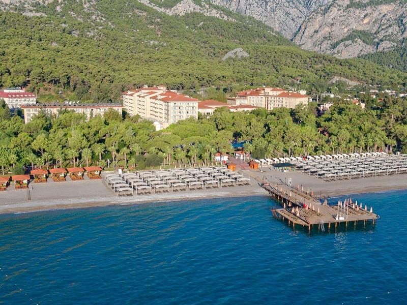 Coastal hotel with beach umbrellas, sunbeds, pier, and green mountains in the background.