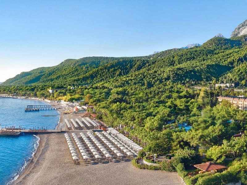 Coastal beach resort with rows of lounge chairs, clear blue sea, and green mountains in the background.