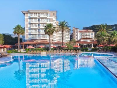 Großer blauer Hotelpool mit Liegestühlen und Palmen, Hintergrund: mehrstöckiges Hotelgebäude.