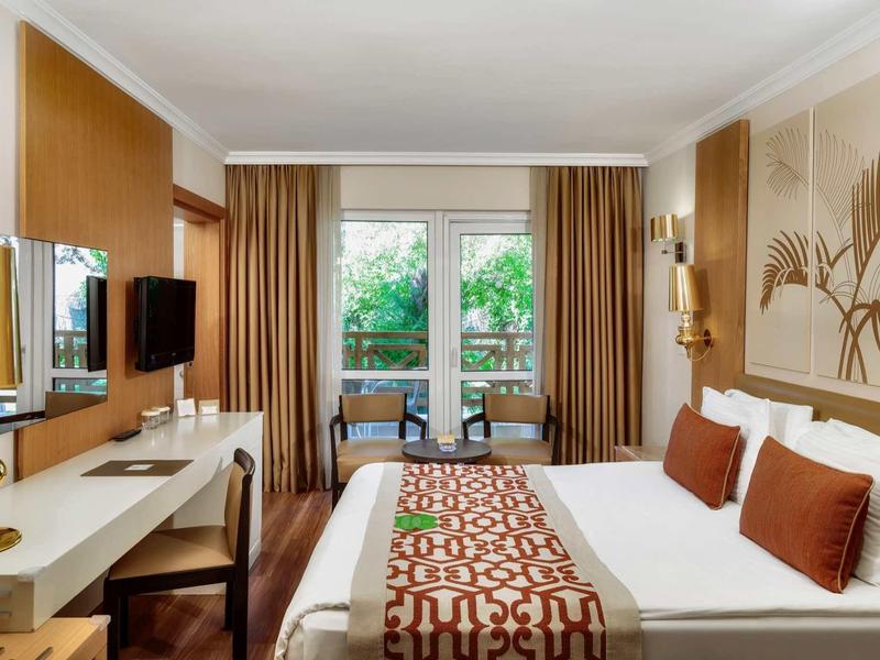 Chambre d'hôtel élégante avec grand lit, décor beige et vue sur jardin.