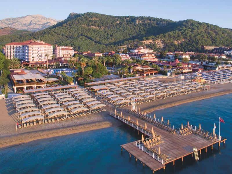 Plage avec parasols et ponton en bois, hôtel et collines à l'arrière-plan.