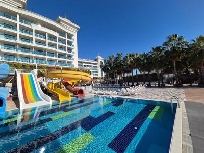 Pool mit Wasserrutschen und Palmen neben einem modernen Hotel bei klarem Himmel.