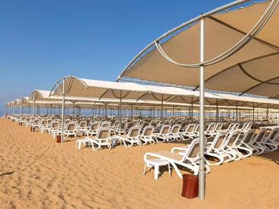 Strand mit Reihen von weißen Liegestühlen und großen Sonnenschirmen auf sandigem Boden.