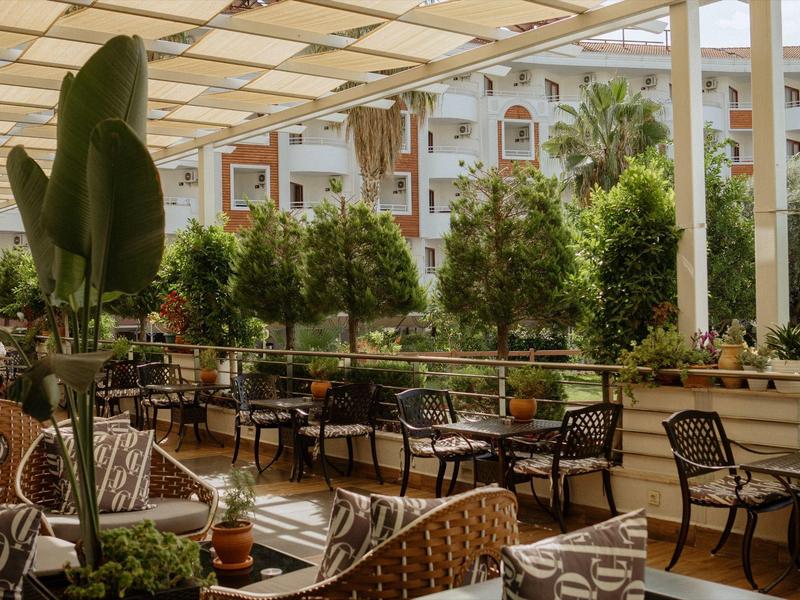 Taras hotelowy z metalowymi stołami, krzesłami i roślinnością pod dachiem z tkaniny.