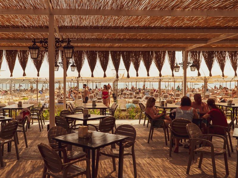 Zadaszony taras kawiarni z drewnianymi stołami i widokiem na zatłoczoną plażę.