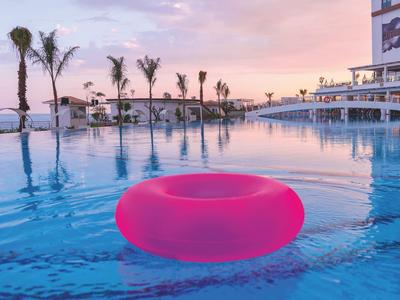 Ciambella gonfiabile rosa che galleggia in una piscina al tramonto con palme sullo sfondo.
