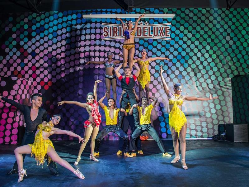 Groupe de danseurs en costumes colorés devant une scène avec le panneau 'Sirius Deluxe'.