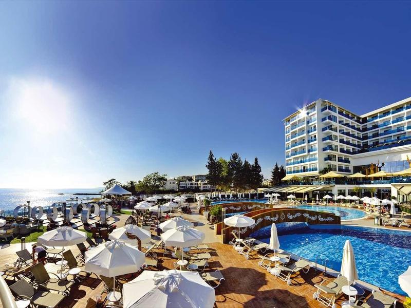 Moderní hotel s venkovním bazénem a lehátky u moře pod slunečnou oblohou.