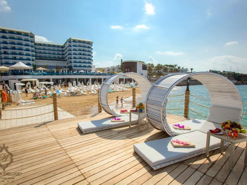 Dřevěná terasa u vody s dvoulůžky a hotelem v pozadí.