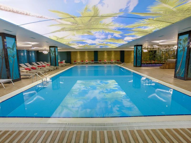 Moderní krytý bazén s osvětleným stropem zobrazujícím modrou oblohu s mraky.