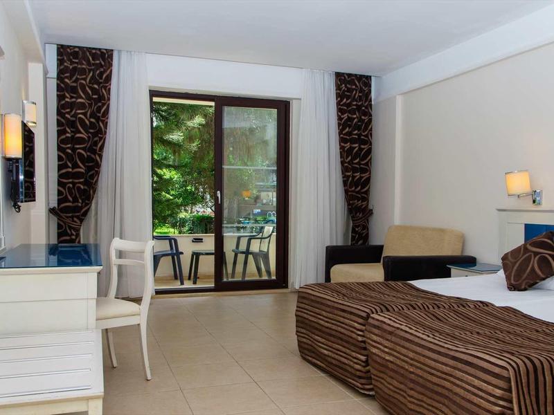 Hotel room with double bed, desk, sofa, and balcony overlooking green trees.