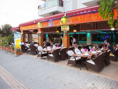 Straßencafé mit geflochtenen Stühlen, vielen sitzenden Gästen und roten Leuchtschildern.