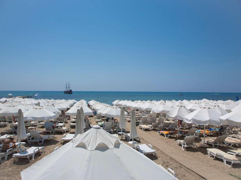 Plaża z wieloma białymi parasolami, leżakami i widokiem na morze z żaglowcem.