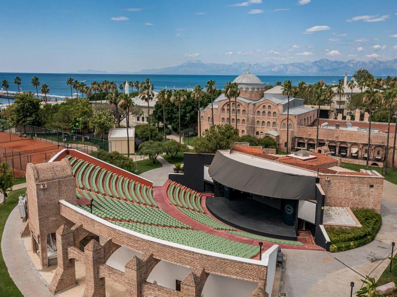 Openluchttheater met halfronde zitplaatsen naast palmbomen en zee op de achtergrond.
