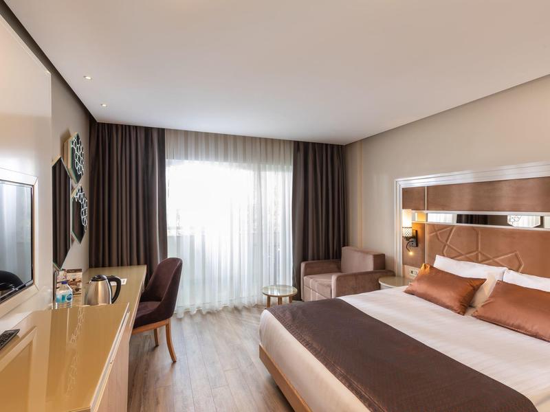 Lichte hotelkamer met een tweepersoonsbed, bureau, stoel en balkonraam met gordijnen.