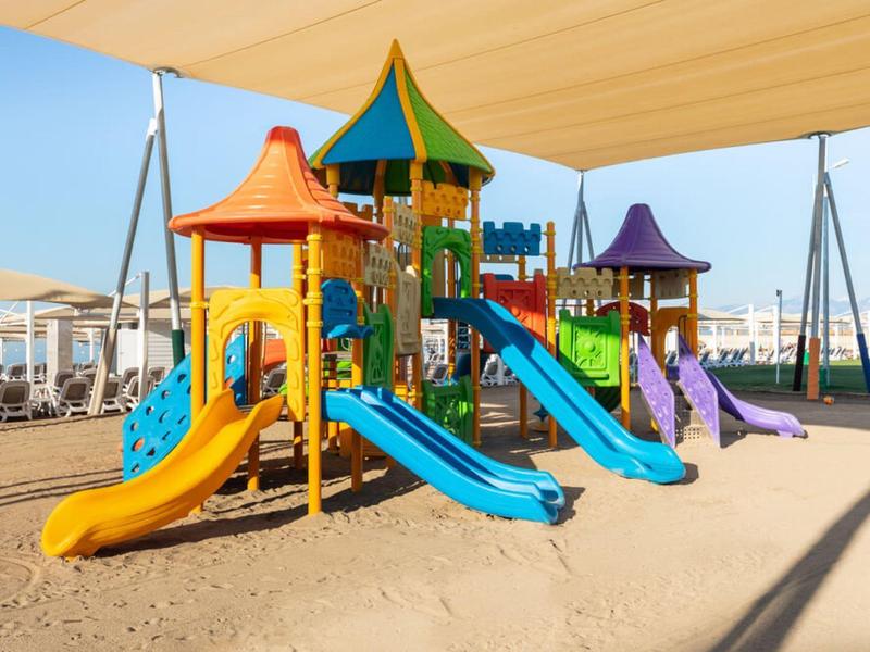 Kleurrijk kinderspeeltoestel met glijbanen onder zonnescherm op zandstrand.