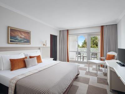 Helles Hotelzimmer mit großem Bett, beigem Teppich und Balkon mit Sitzgelegenheit.