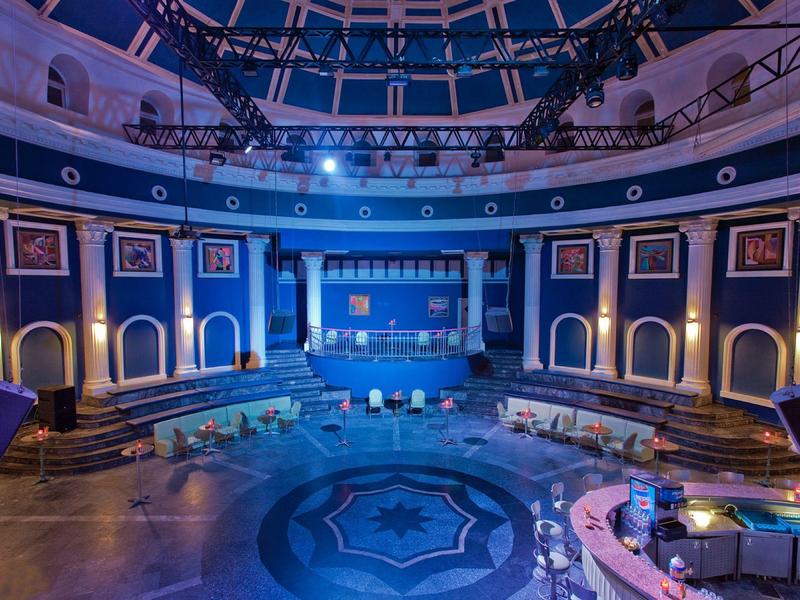 Rundes Inneres eines Theaters mit blauer Beleuchtung, Sitzreihen, Kunstwerken an den Wänden und Sternmuster am Boden.