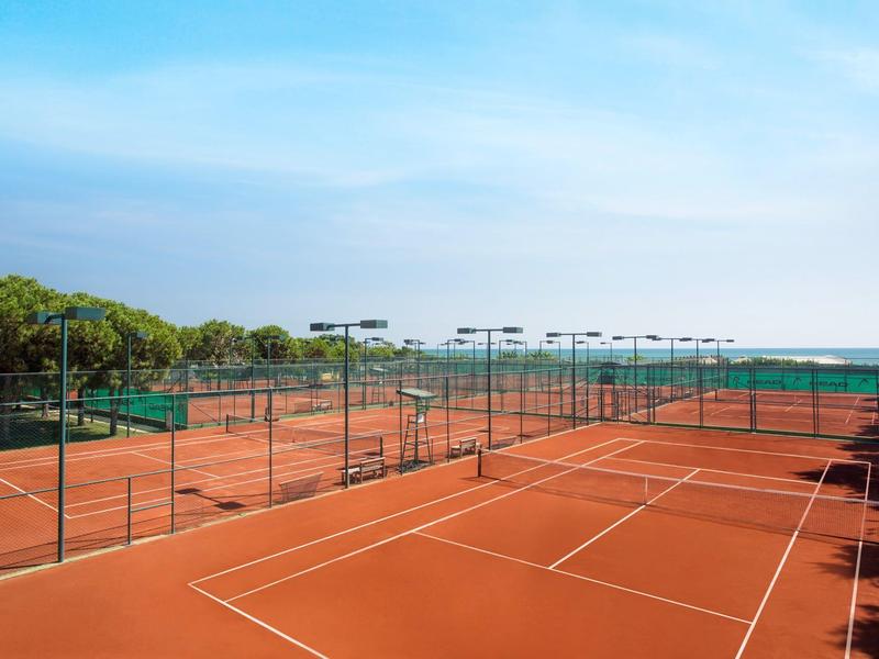 Mehrere rote Sand-Tennisplätze mit grünen Zäunen unter blauem Himmel.
