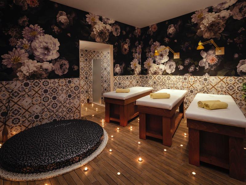 Gemütlicher Spa-Raum mit Blumenmustertapeten, zwei Massageliegen, Kerzenlicht und großer runder Matte.