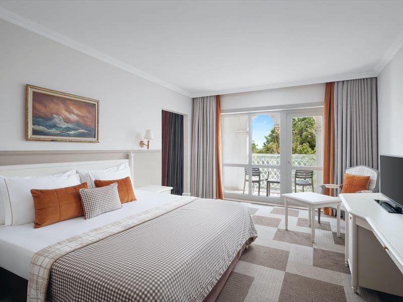 Helles Hotelzimmer mit großem Bett, beigem Teppich und Balkon mit Sitzgelegenheit.