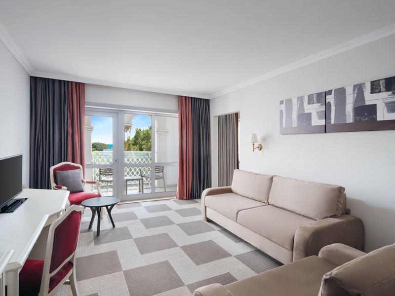 Helles Hotelzimmer mit Sofa, Sesseln, Fernseher und Balkon mit Blick ins Grüne.