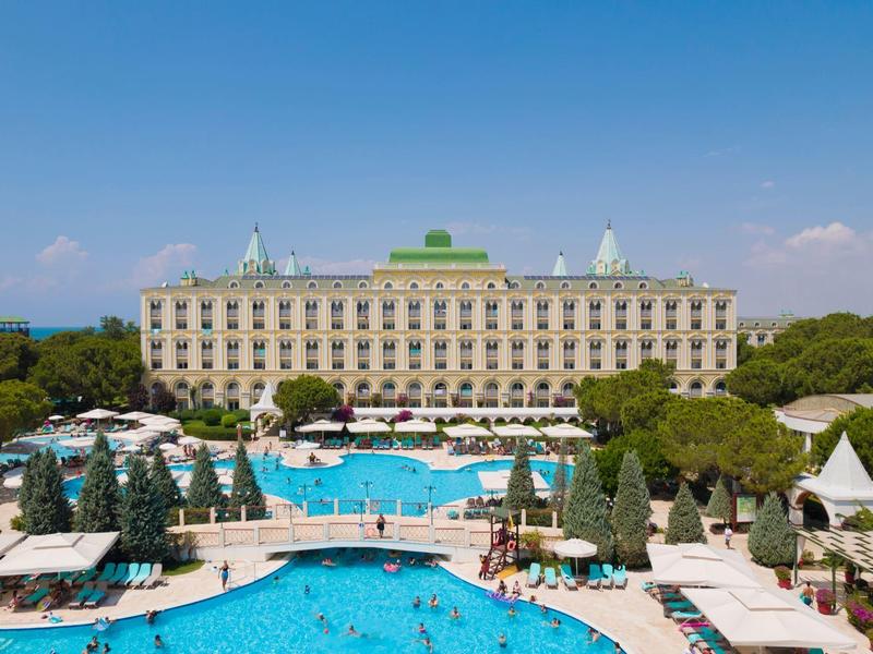 Großes Hotel mit blauem Außenpool und Sonnenliegen in einer sonnigen Umgebung.